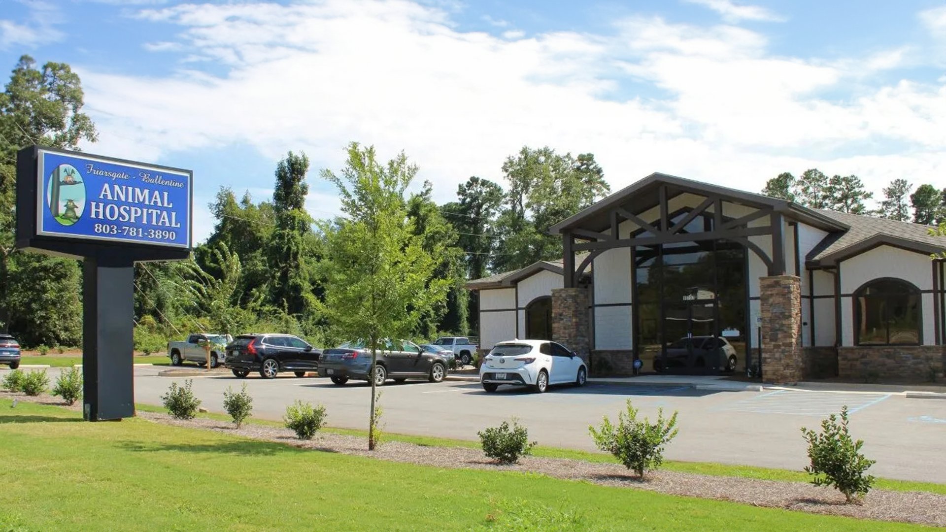 The exterior of Friarsgate-Ballentine Animal Hospital, showing a modern building with a large entrance featuring stone pillars and dark wooden beams. Several cars are parked in the parking lot in front of the building. A sign on the left reads "Friarsgate-Ballentine Animal Hospital" with the phone number 803-781-3890. The surrounding area has neatly landscaped greenery and trees.