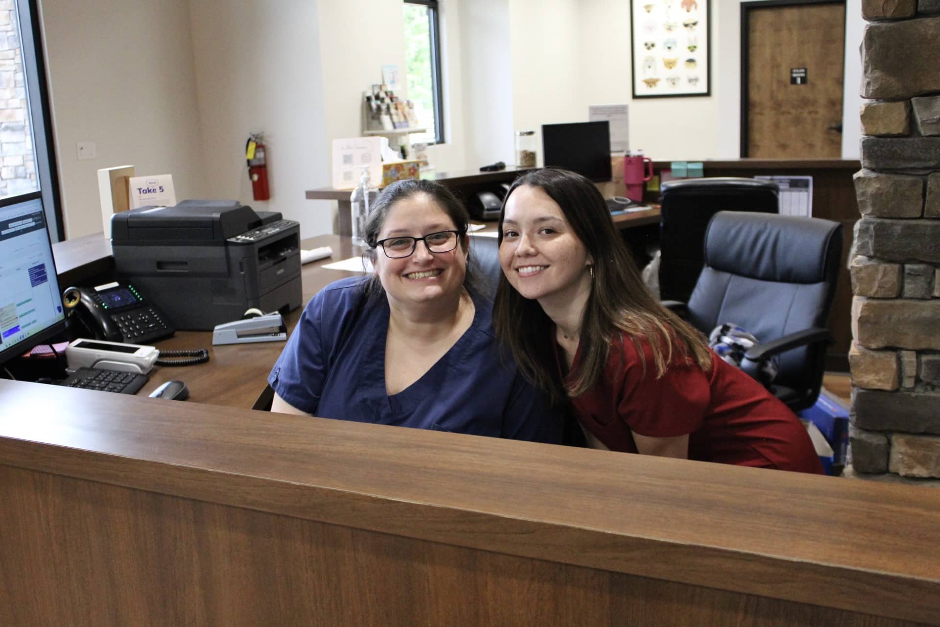 Two smiling staff members at Friarsgate-Ballentine Animal Hospital are seated at the reception desk. The person on the left is wearing navy scrubs and glasses, while the person on the right is wearing maroon scrubs. The reception area is equipped with a computer, printer, and other office supplies. The background features a window, some decor, and a doorway leading to another room. Both individuals appear friendly and welcoming, adding a warm atmosphere to the hospital.